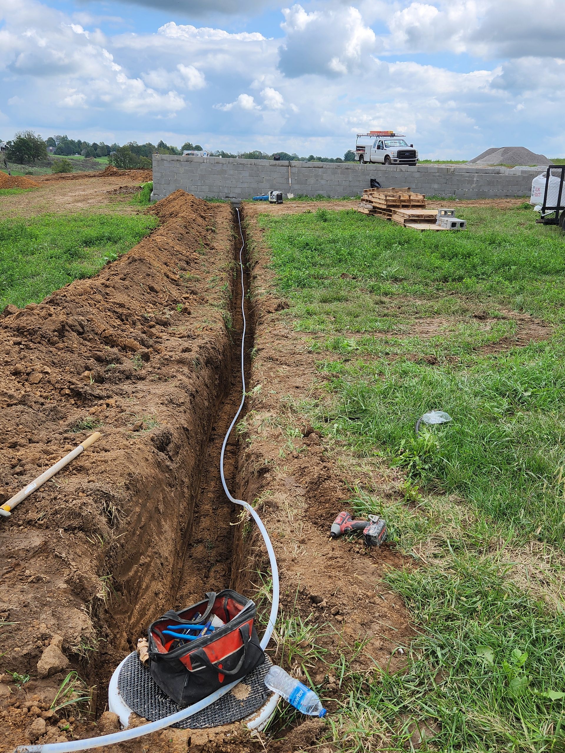 Trench with exposed white pipe and cable in a grassy construction site under cloudy sky