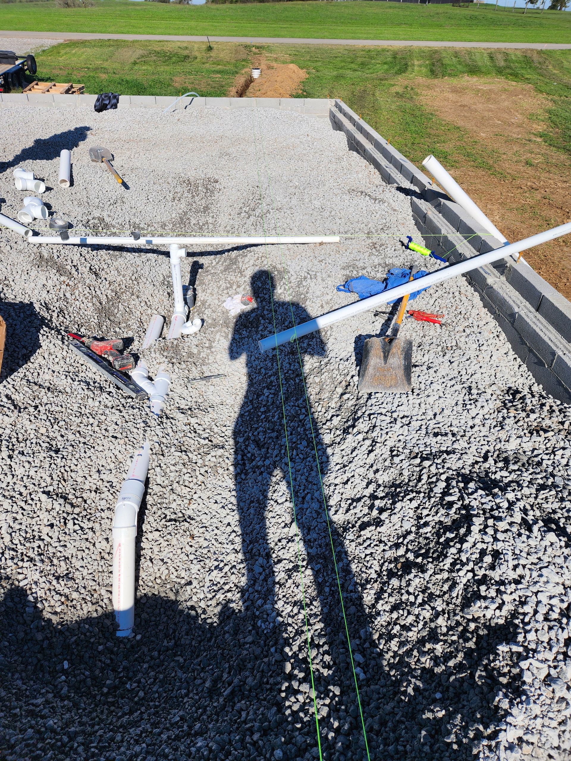Shovel stands in a gravel trench with white PVC pipes and a blue-handled tool on a sunny lawn.