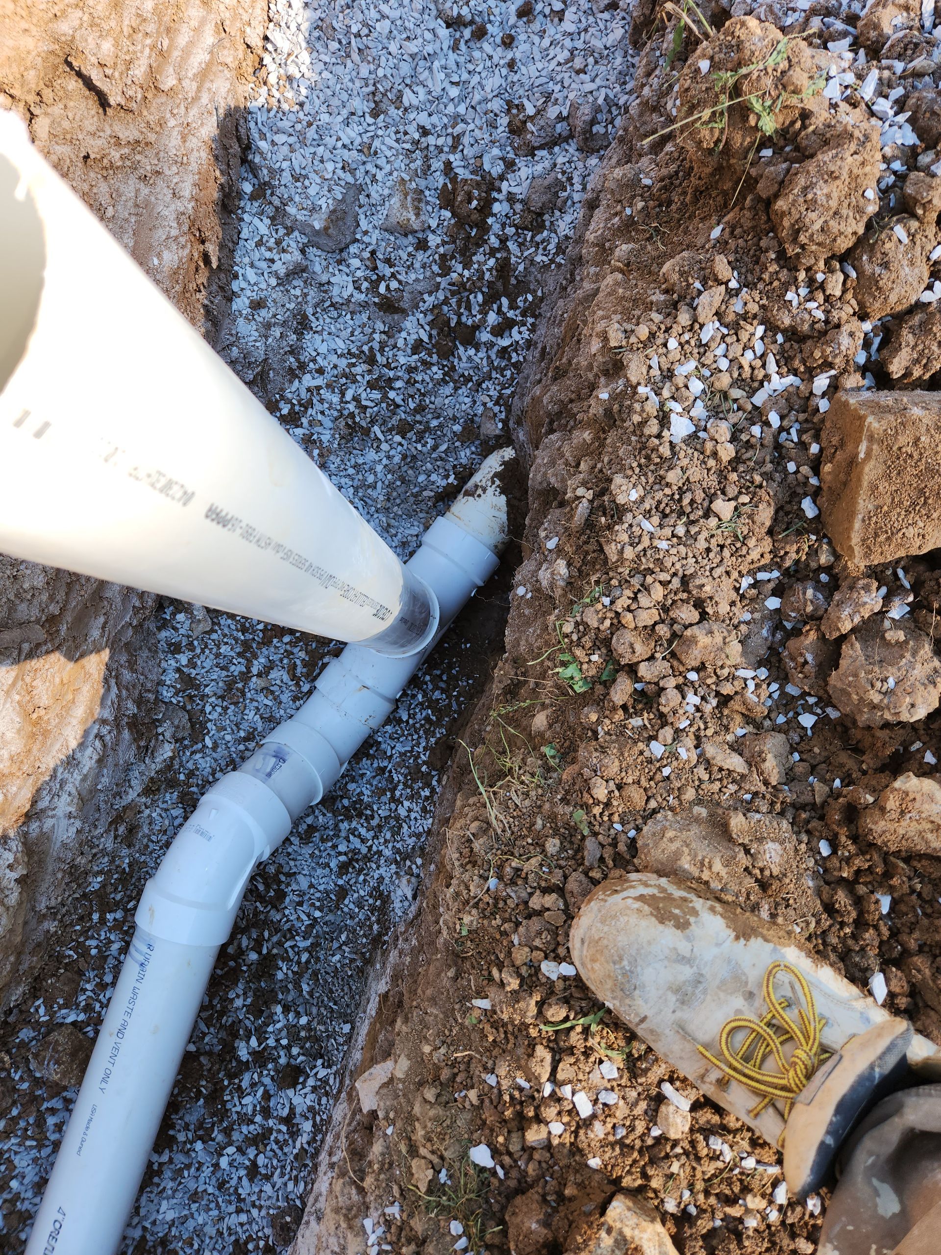 White PVC pipe in a trench beside gravel and dirt, with a worker’s boot visible nearby.