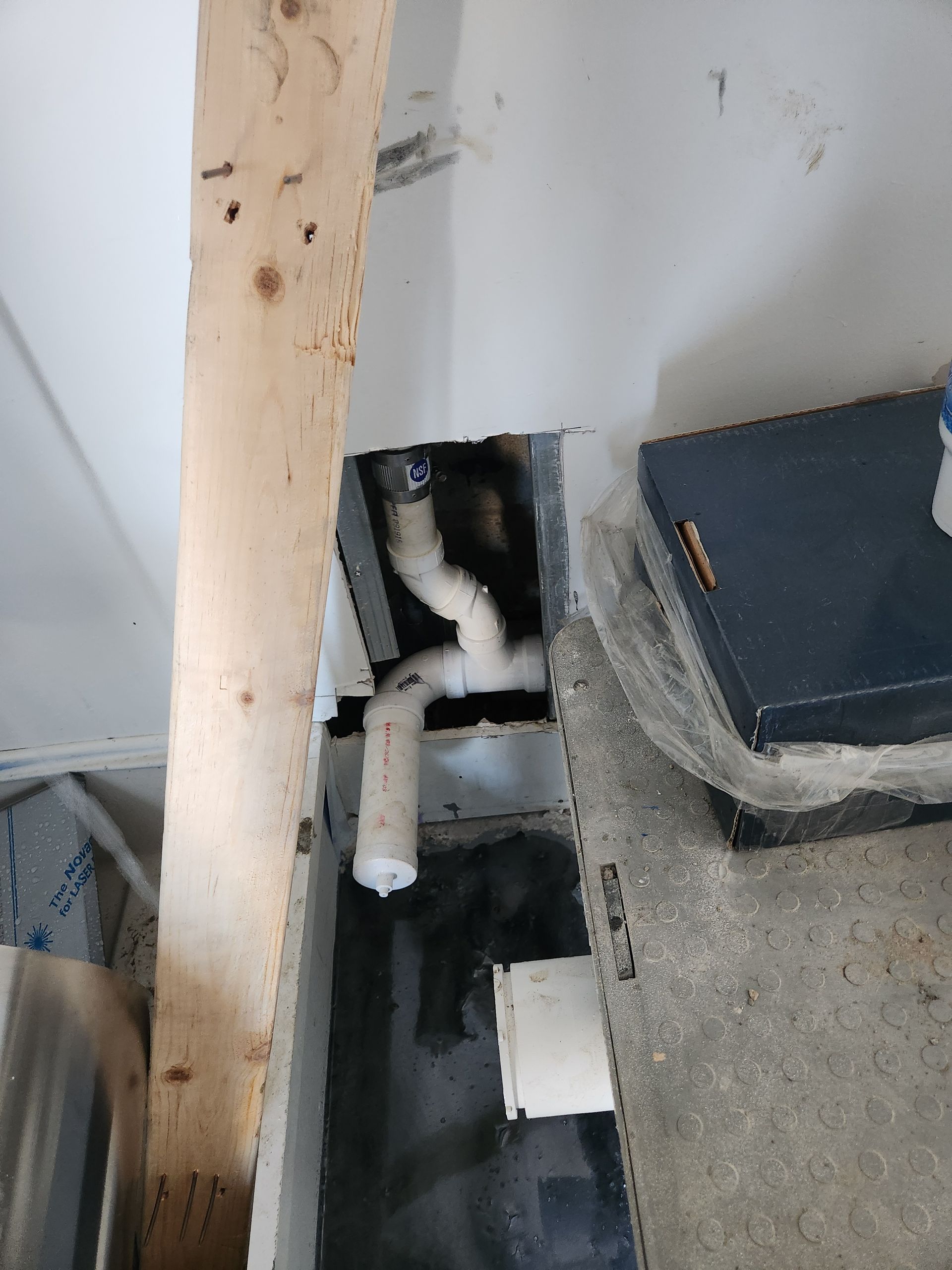 Black and white cat peeking from a gap beside pipes and a blue box on the floor