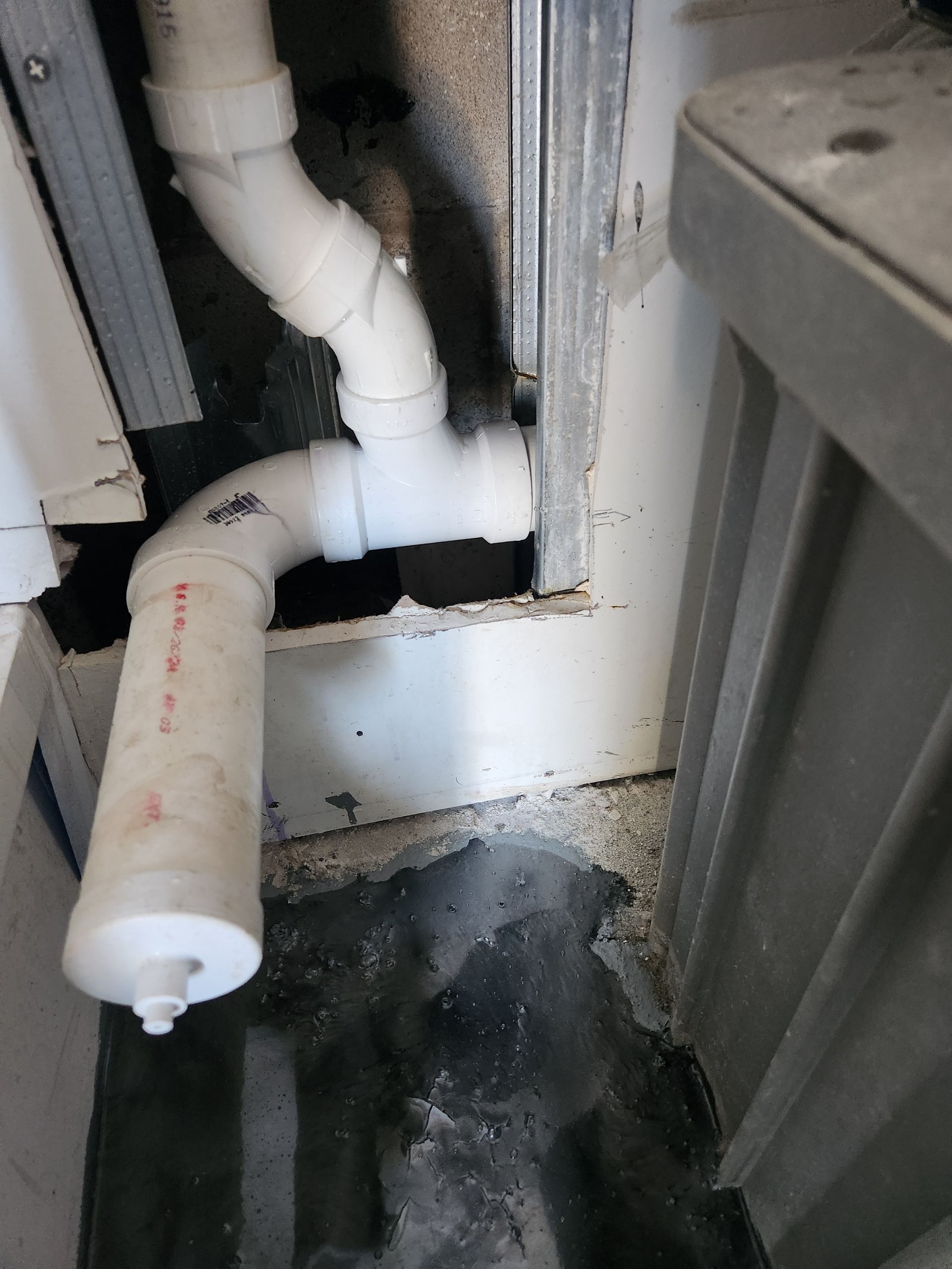 White plumbing pipes under a sink beside a gray cabinet, with water damage and dark mold stains on the floor.