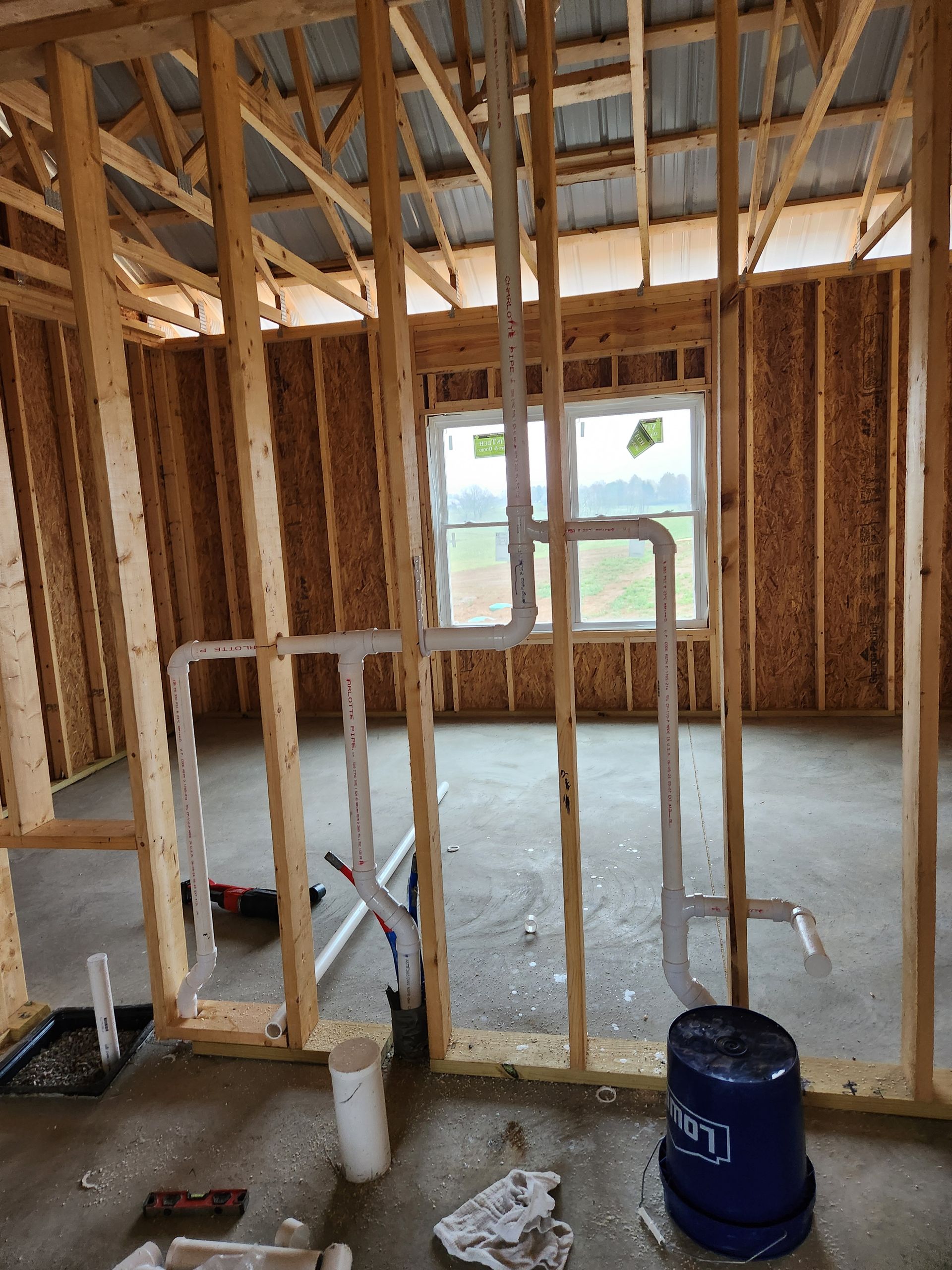 Unfinished room with exposed wooden studs, insulation, concrete floor, and a blue bucket near a window.