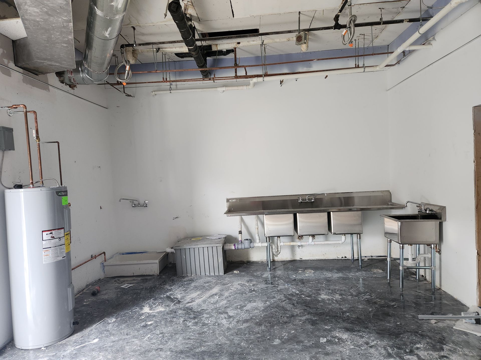 Empty industrial room with a stainless worktable, utility sink, water heater, and unfinished ceiling.