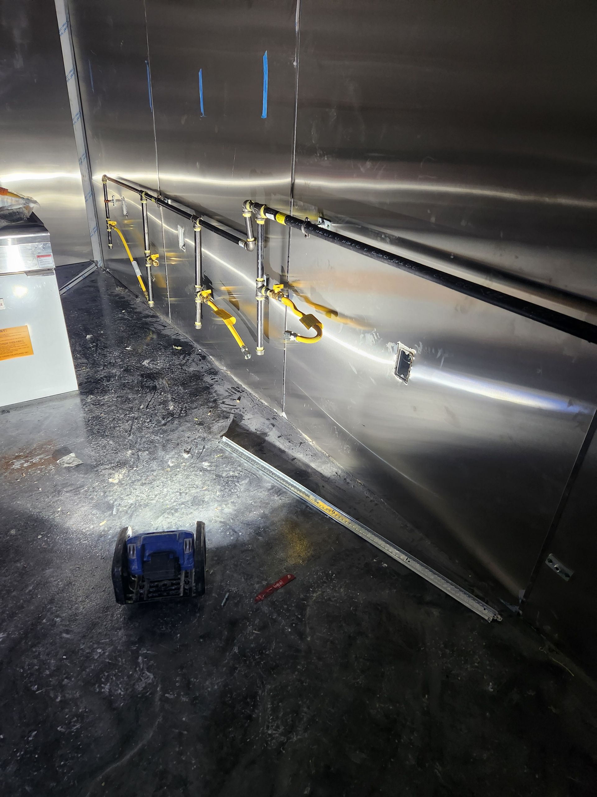 Metal wall in a service corridor with yellow pipes and a small blue device on the floor