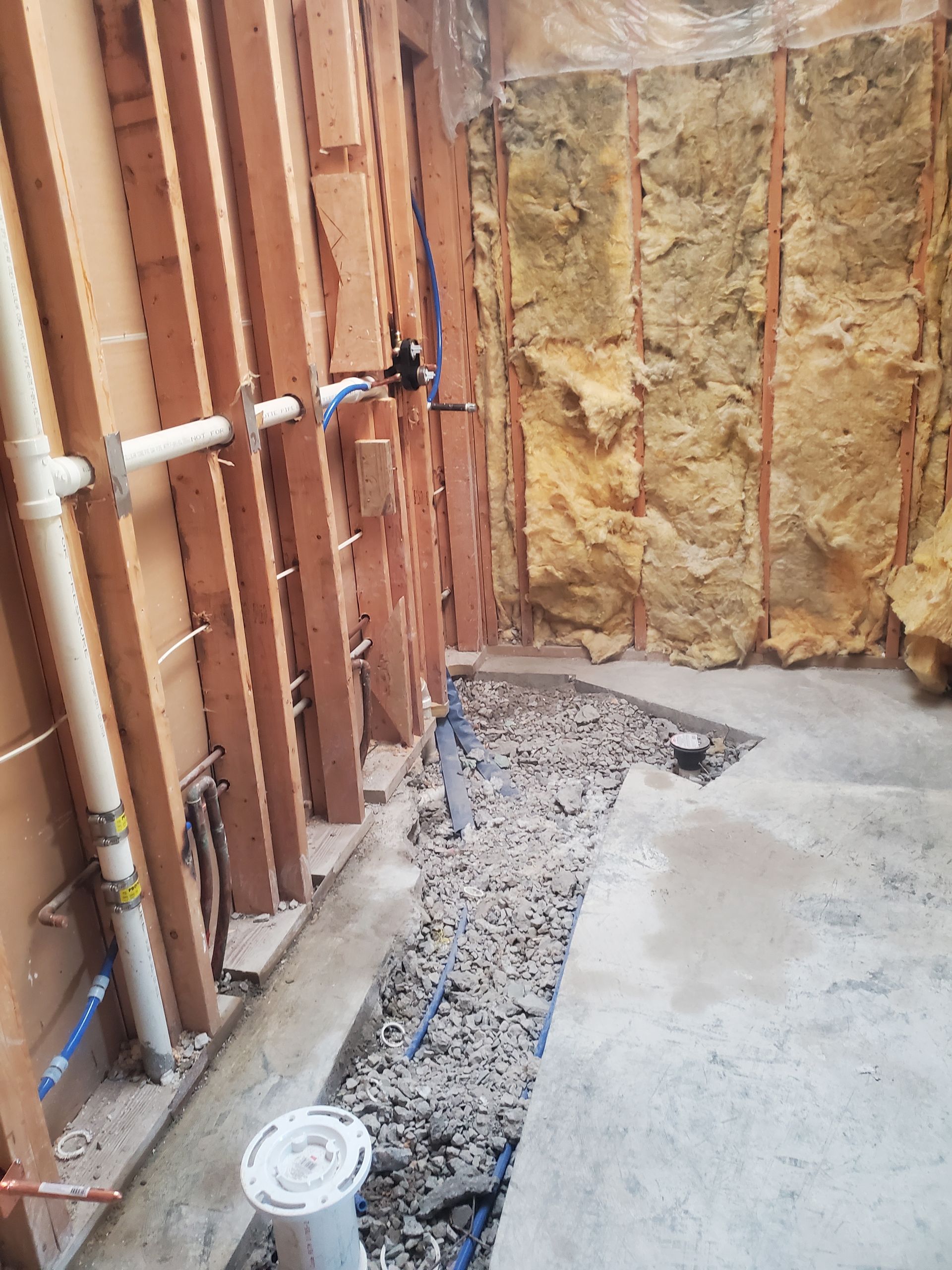 Construction trench beside wall framing and insulation, with exposed plumbing pipes and gravel floor.