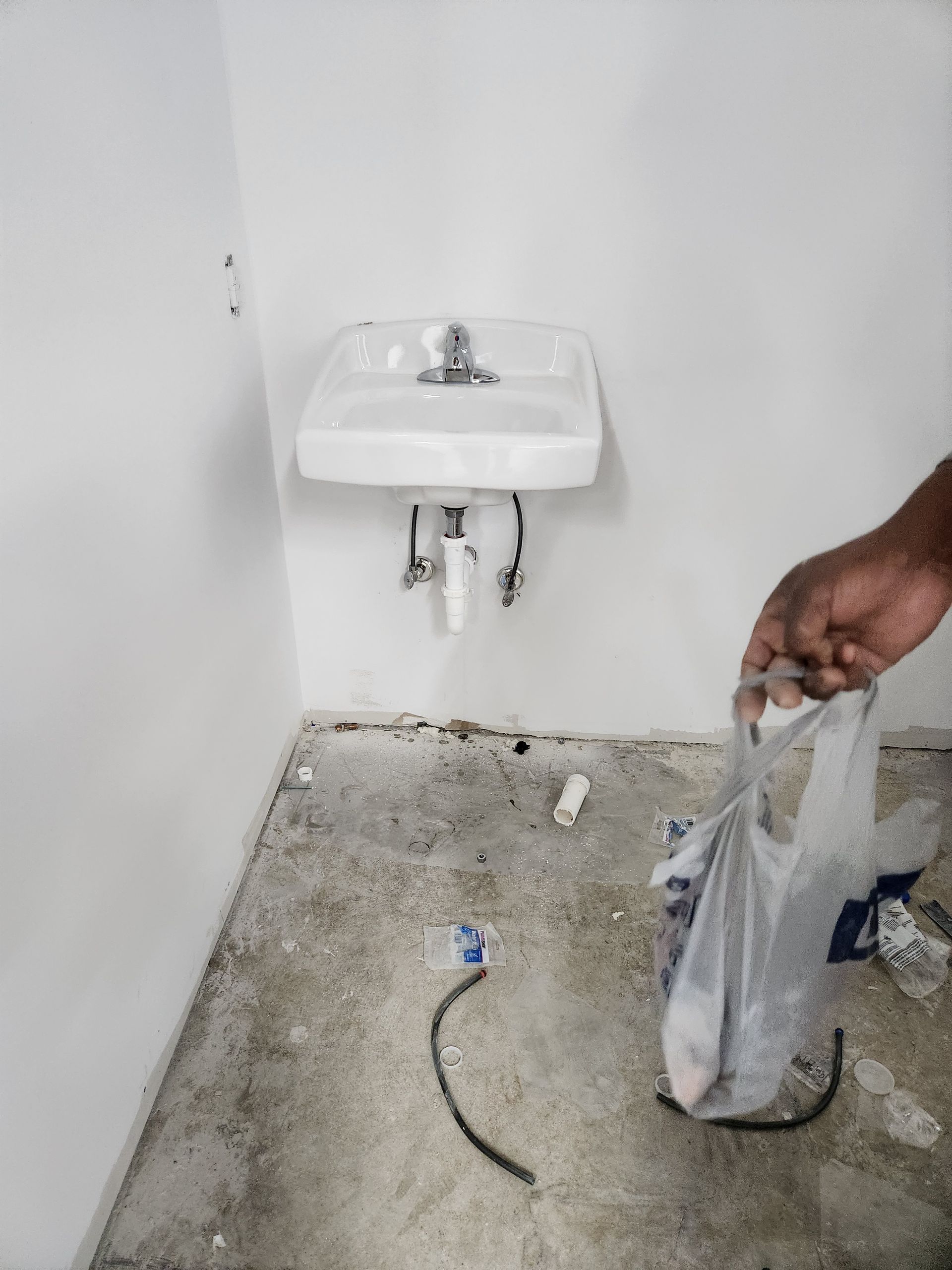Small sink on white wall in unfinished room, with a person holding a plastic bag near exposed plumbing.
