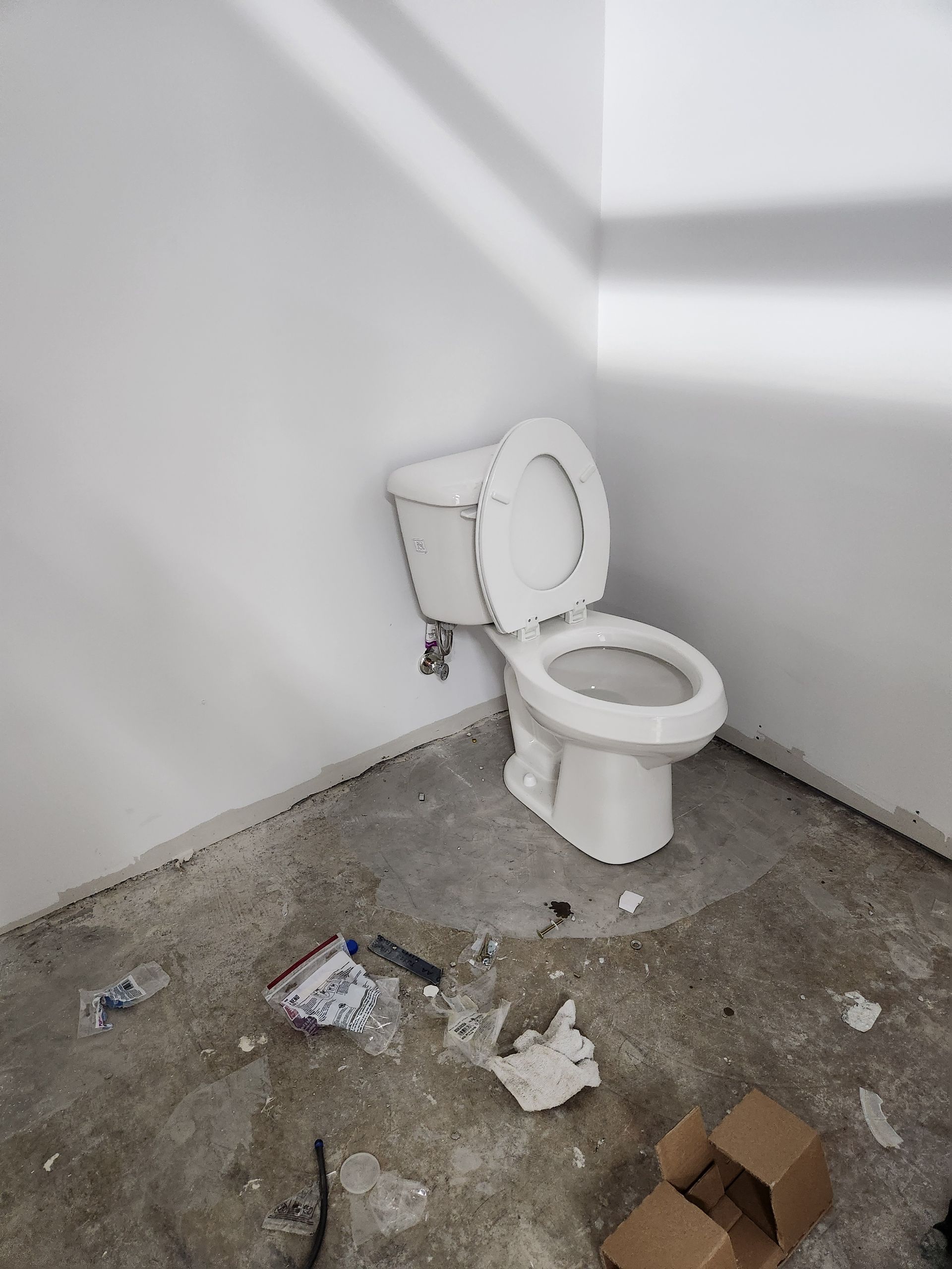 Small unfinished bathroom with a toilet, stained concrete floor, and scattered debris and cardboard.
