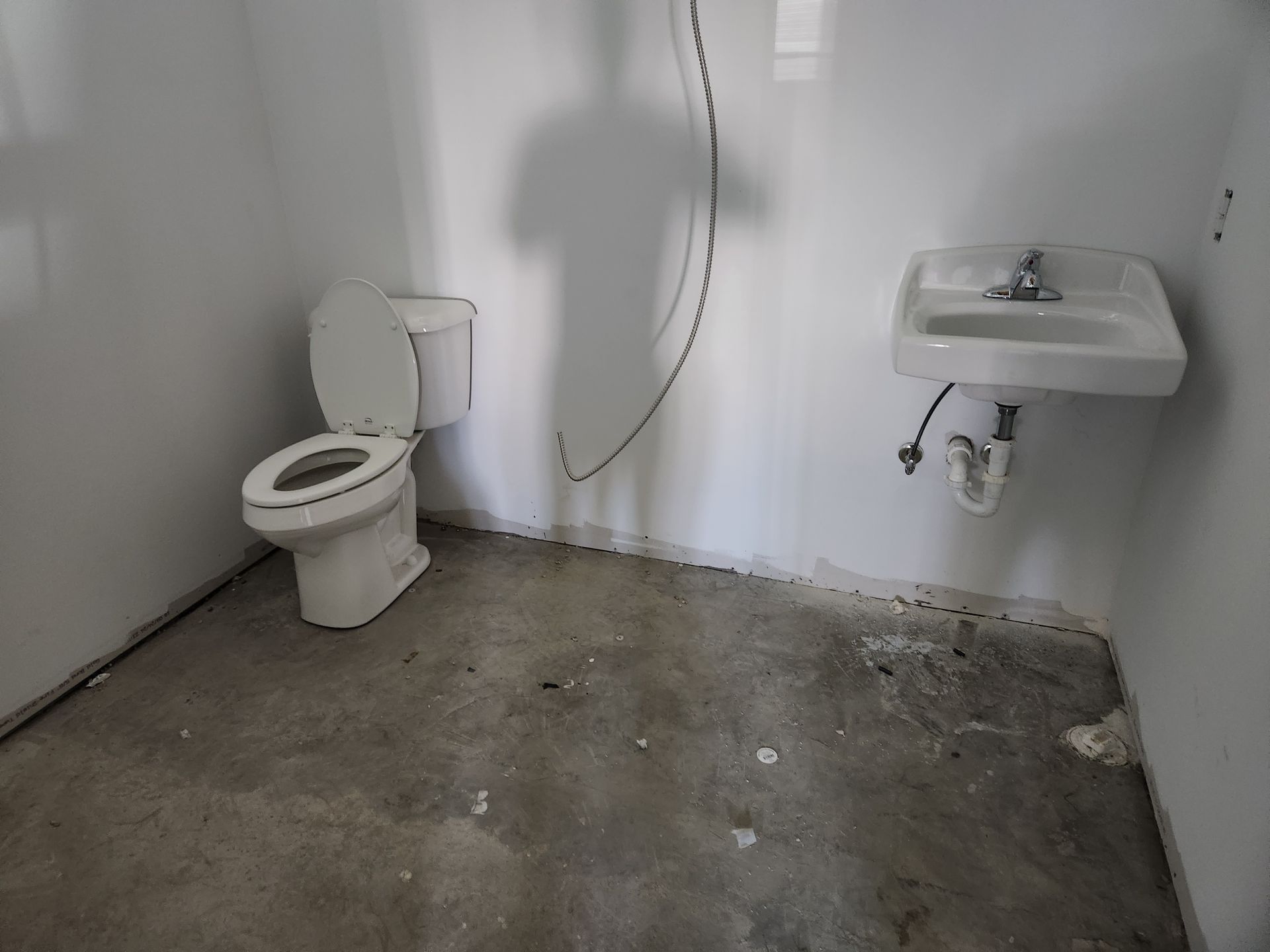 Small bare bathroom with toilet, wall-mounted sink, exposed shower hose, and concrete floor.