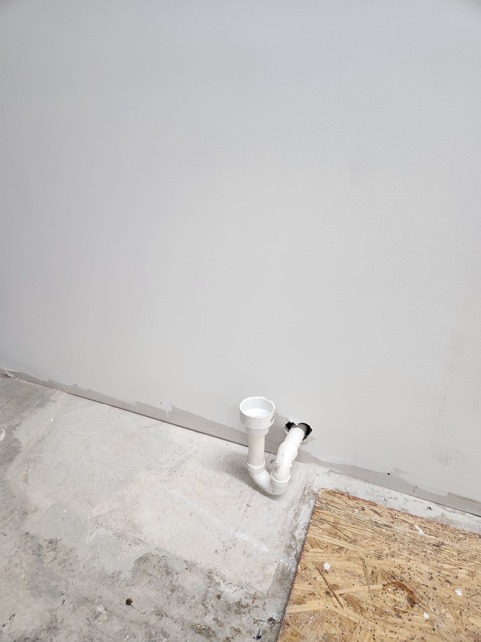 White PVC plumbing pipes protruding from a wall above a concrete floor and plywood subfloor during construction.