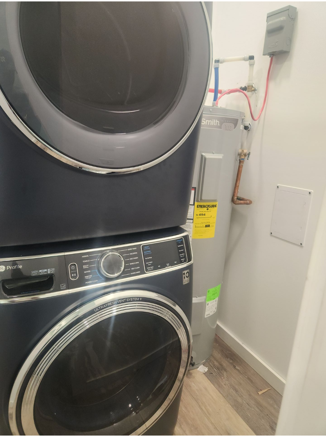 Stacked black washer and dryer in a narrow laundry room beside a water heater and wall pipes.