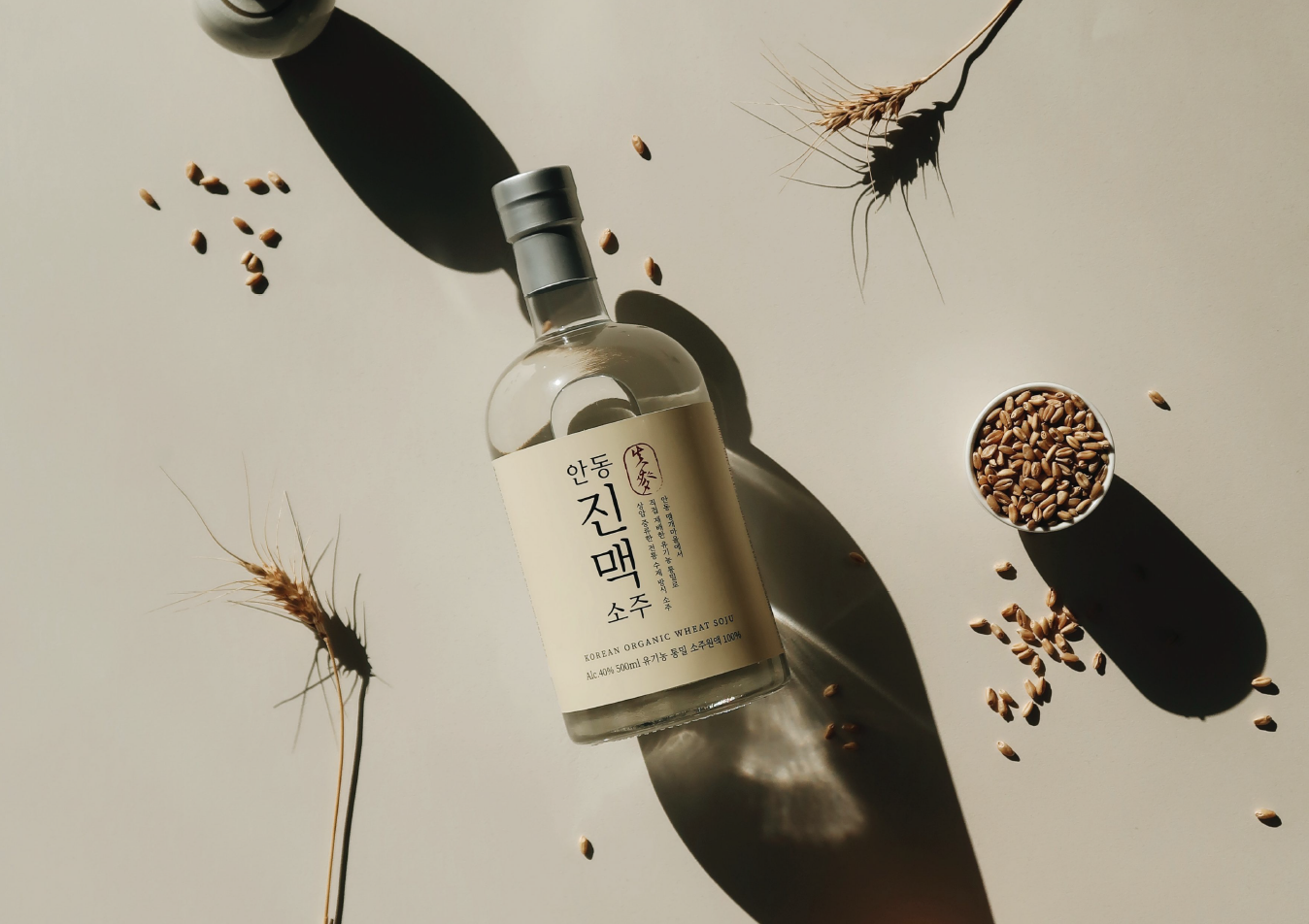 베이지색 표면 위에 한국 소주가 담긴 투명한 유리병이 놓여 있고, 그 주변에 곡물과 말린 밀 줄기가 놓여 있습니다.