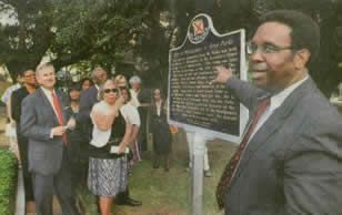 Man in suit points to a historical marker while others stand nearby outdoors.