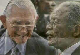 Two elderly men in suits speaking face-to-face outdoors, one smiling warmly