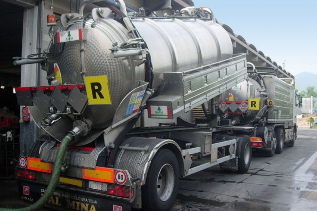 Autocisterne che trasportano grandi serbatoi industriali argentati parcheggiate in un piazzale, con cartelli di avvertimento per materiali pericolosi ben visibili.