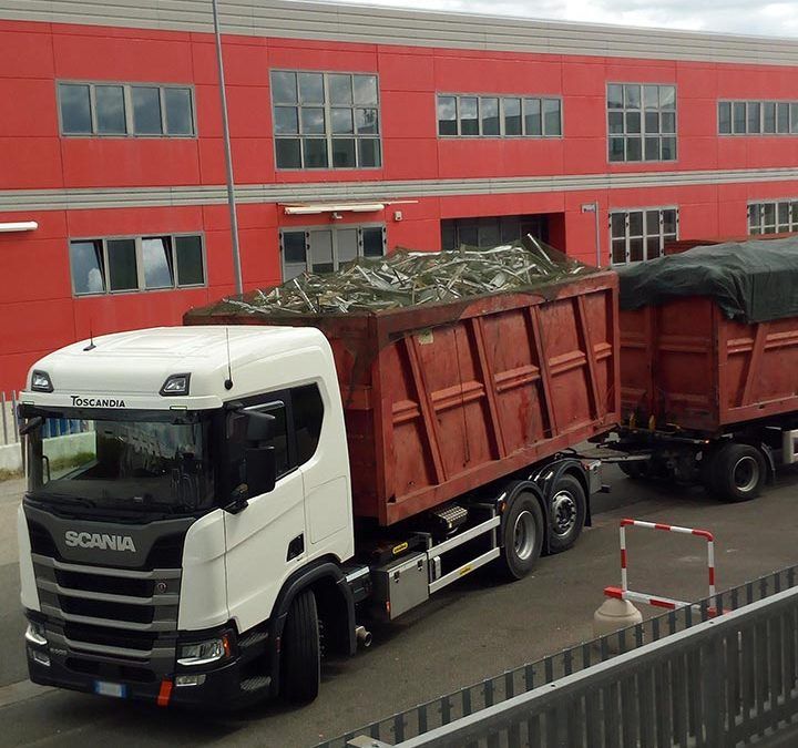 Un camion Scania bianco con rimorchio rosso carico di detriti è parcheggiato accanto a un edificio industriale rosso.