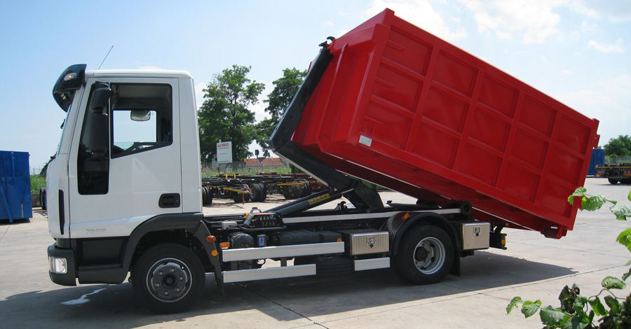 Un camion bianco solleva un cassonetto rosso in un cantiere.