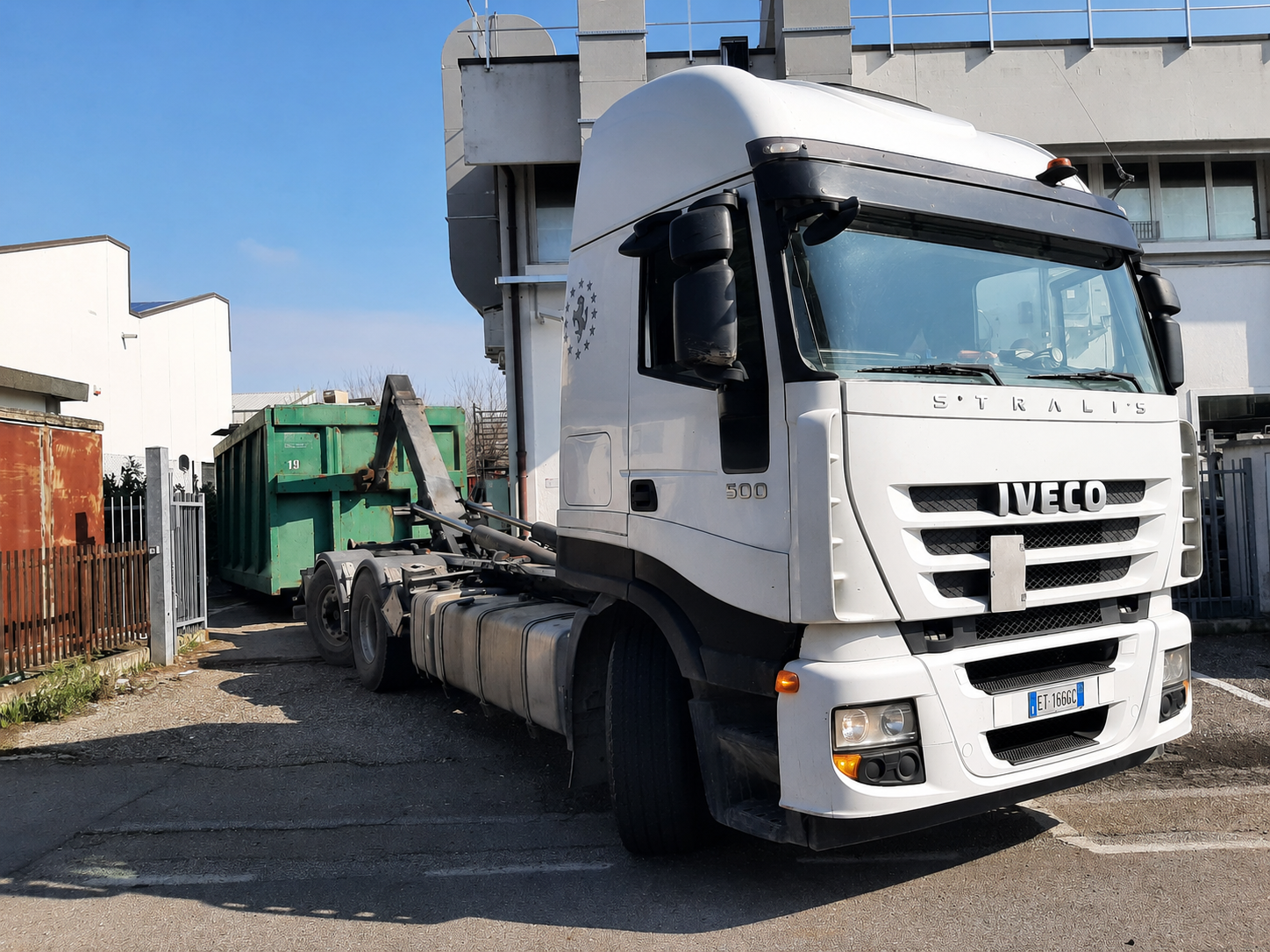 Un camion Iveco bianco è parcheggiato accanto a un cassonetto verde in un'area industriale.