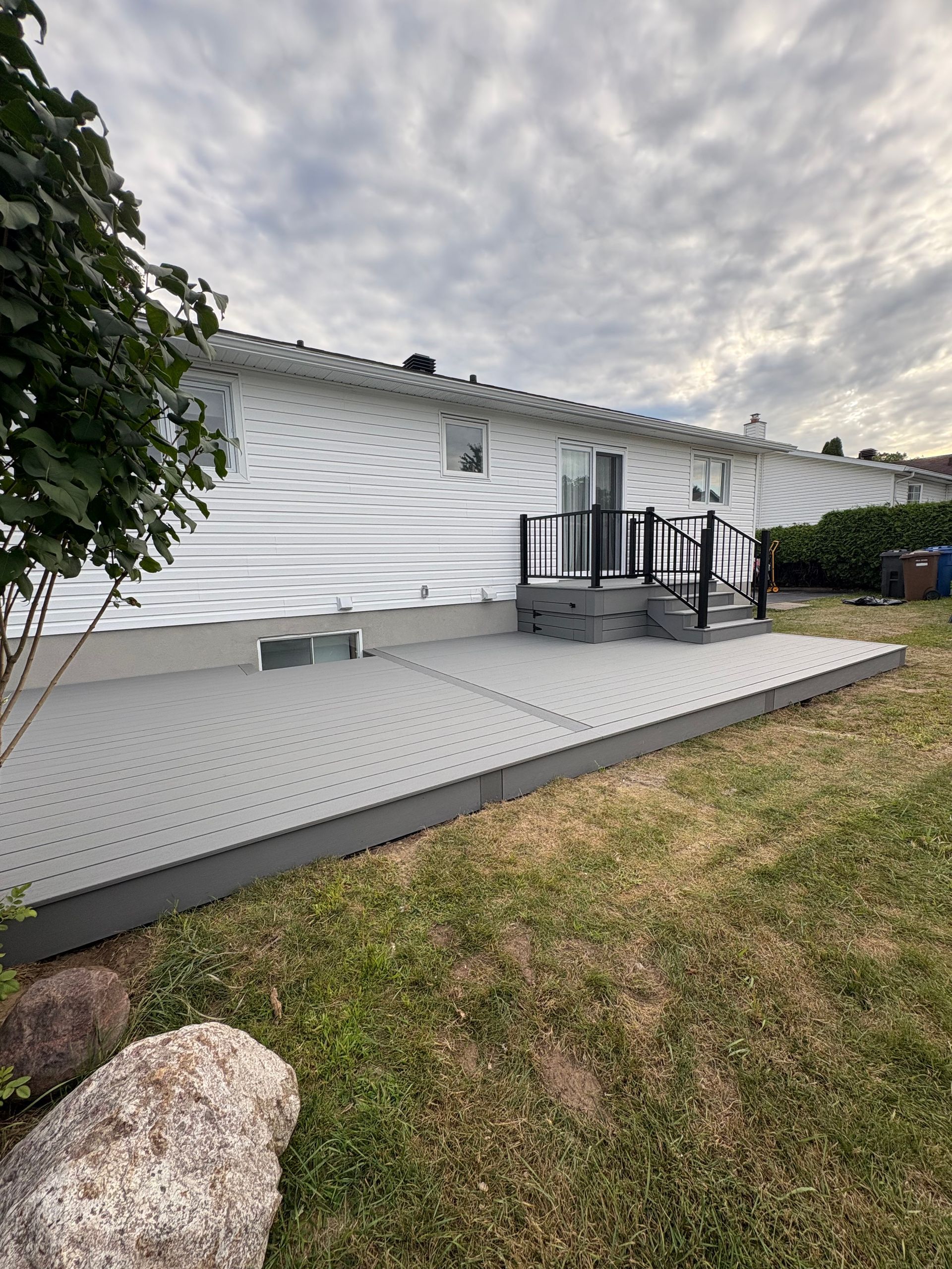 Terrasse en composite gris avec marches, rambarde noire, devant une maison blanche, ciel nuageux.