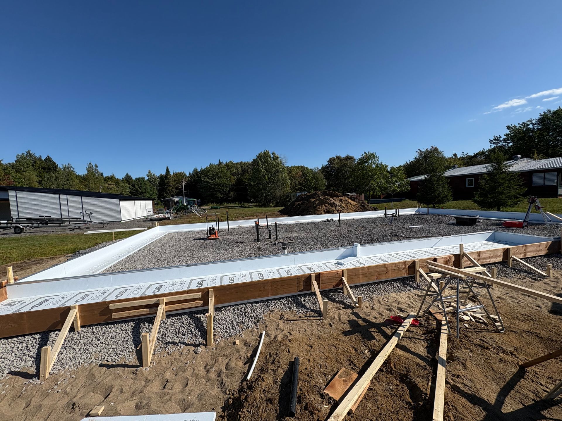 Chantier avec coffrages en bois et base de gravier, prêt pour les fondations en béton. Tuyaux apparents. Ciel bleu.