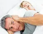 Uma pessoa permanece acordada na cama, tapando os ouvidos com a mão enquanto seu parceiro ronca por perto.