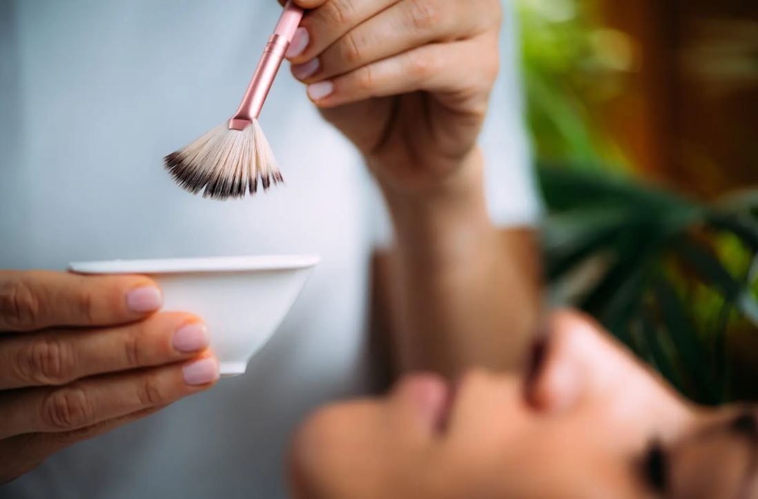 Hands mix a white bowl with a small whisk over a blurred face lying down in a spa setting