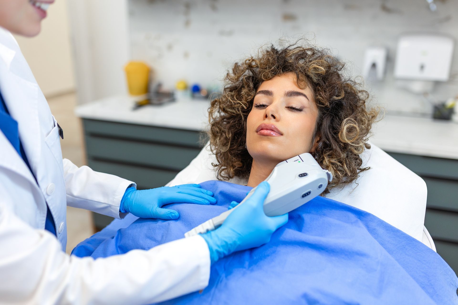Clinician uses a handheld device on a patient covered with a blue blanket in a medical exam room