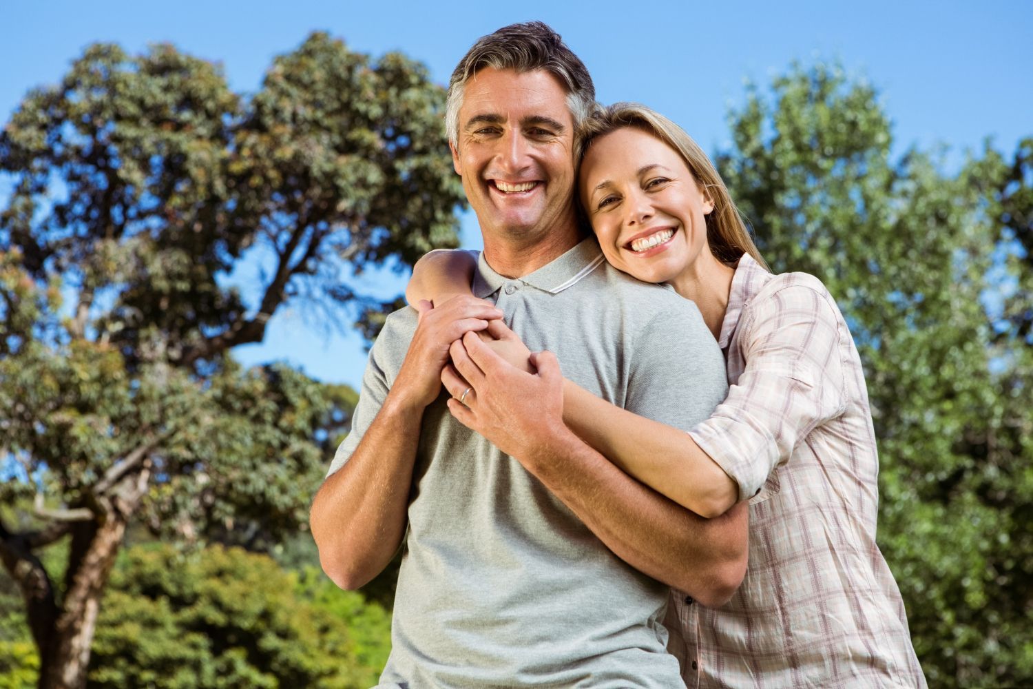 Smiling couple standing together outdoors in a sunny park with trees and blue sky.