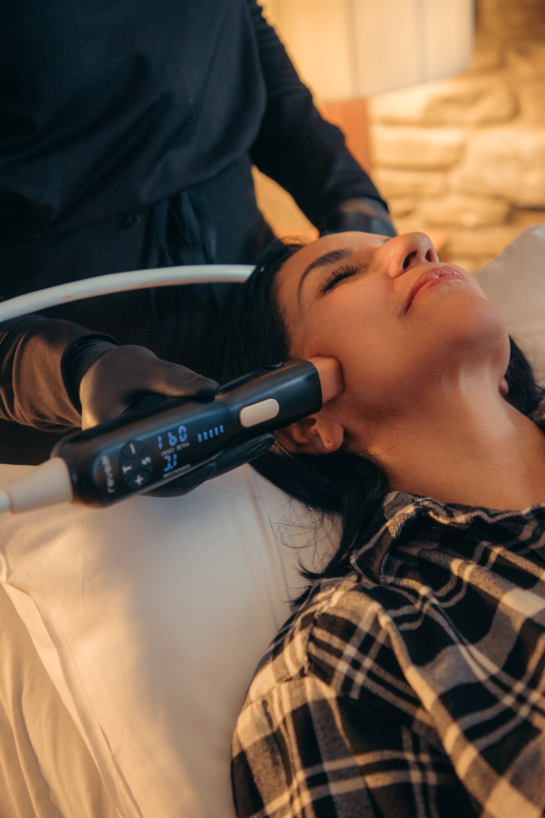 Woman receiving a laser skin tightening treatment. A person in black gloves holds a device to her cheek. Indoors, neutral lighting.