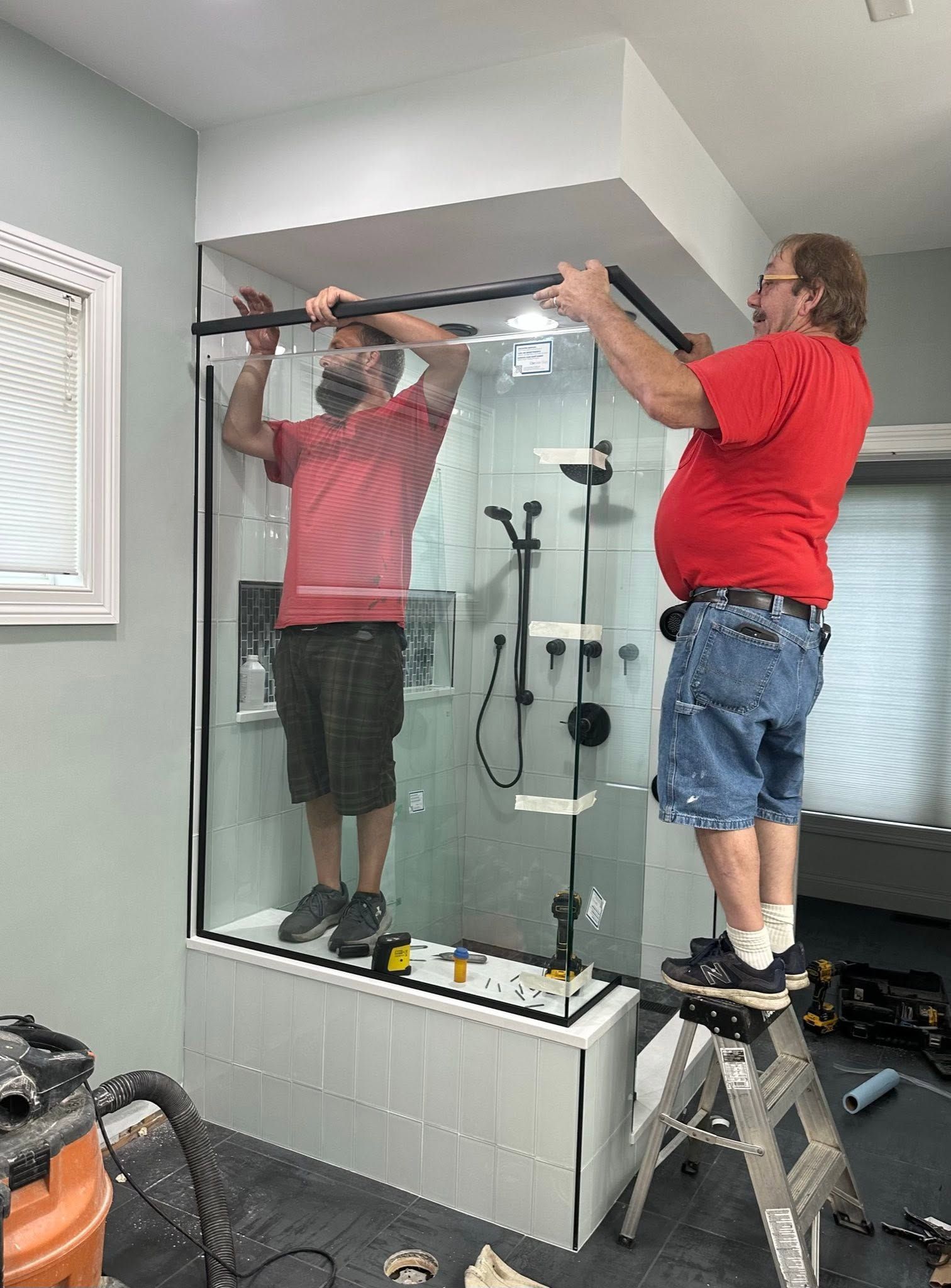 Two workers install a shower enclosure, standing on a ladder and tub in a bathroom.
