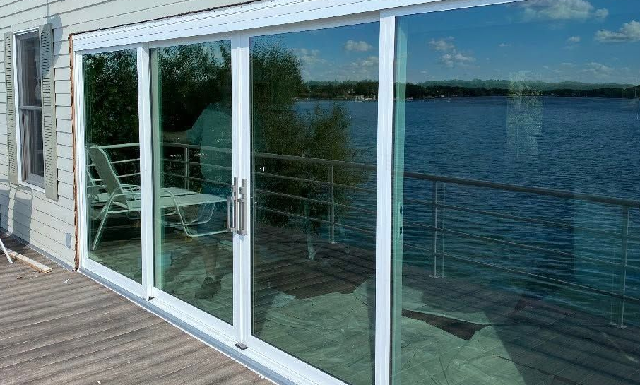 Sliding glass doors on a deck overlooking a calm blue lake, with reflections of patio furniture.