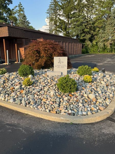 Rock garden with a stone sign in front of a brown building and evergreen trees.