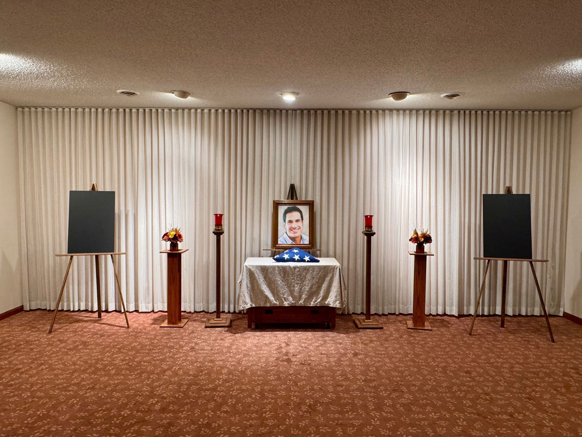 Memorial altar with framed portrait, candles, flowers, and two easels in a curtained room