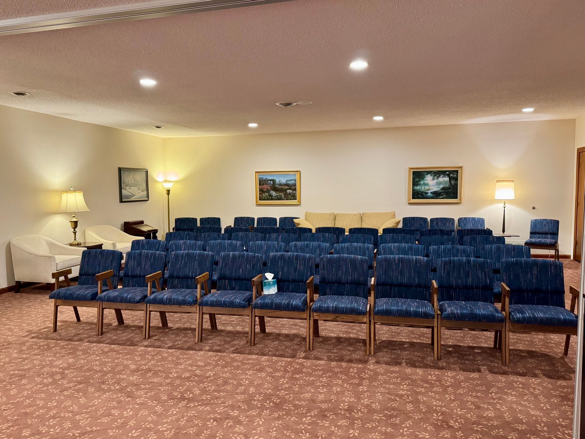 Waiting room with rows of blue chairs, lamps, and framed paintings on beige walls