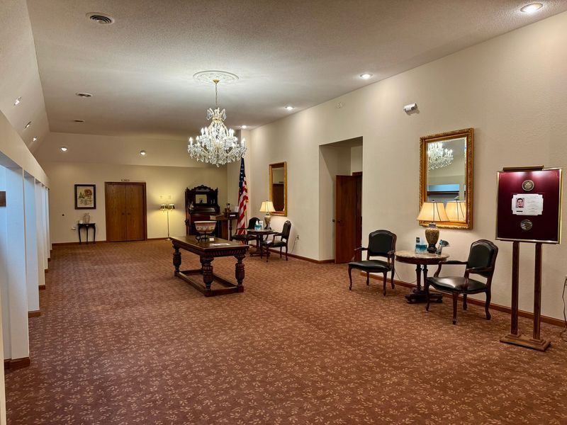 Spacious hotel lobby with patterned carpet, chandelier, chairs, and framed artwork on beige walls