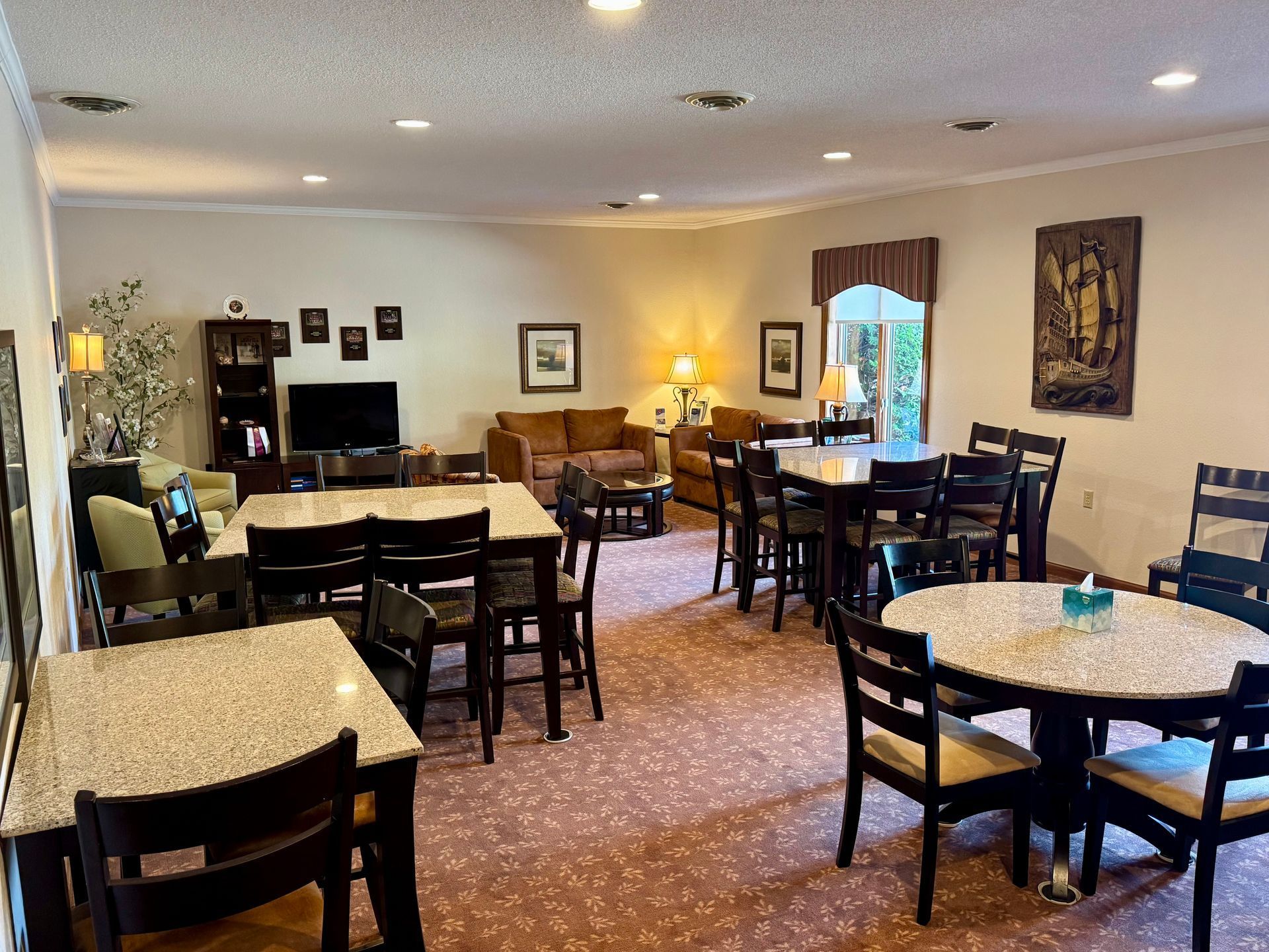 Empty dining room with round and square tables, dark chairs, patterned carpet, and warm ceiling lights.