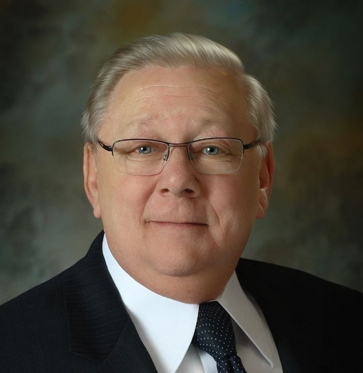 Studio portrait of a man in a dark suit and tie, wearing glasses, against a soft gray background