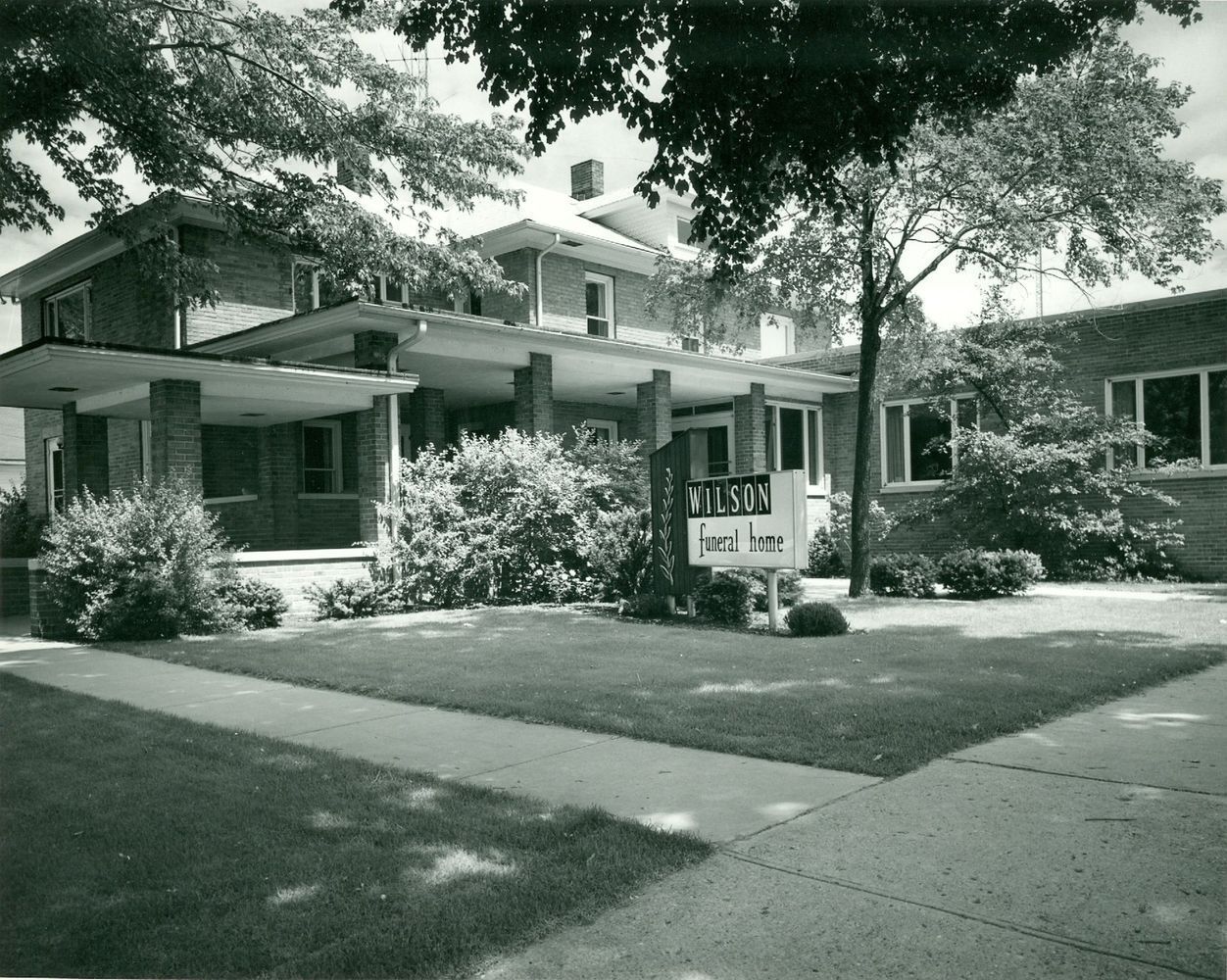Black-and-white photo of a house with a front porch, trees, and a lawn with a sidewalk sign.