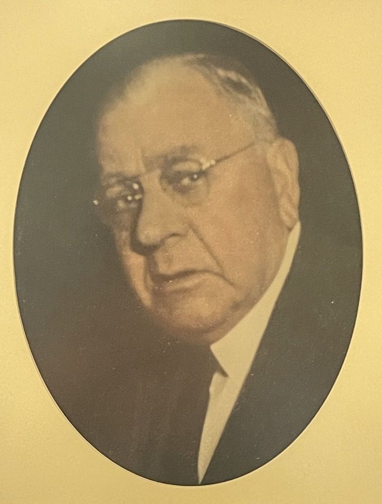 Oval-framed formal portrait of a man in a dark suit and tie against a warm beige background