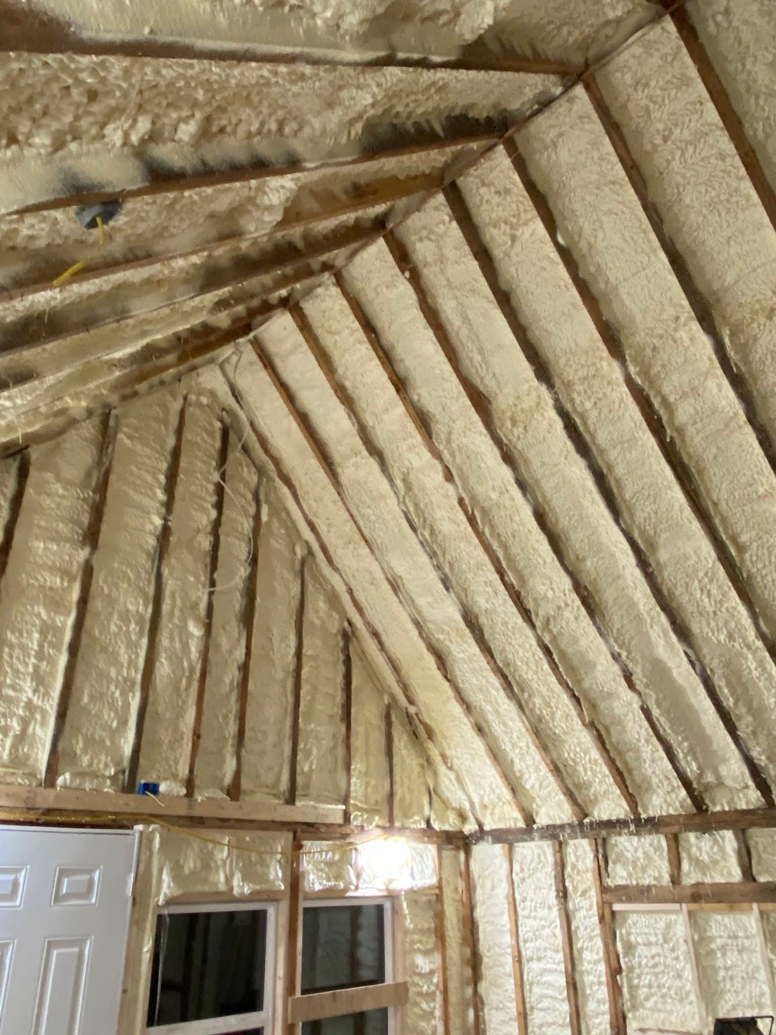 Exposed foam-insulated ceiling framing above a room with white doors and windows below.
