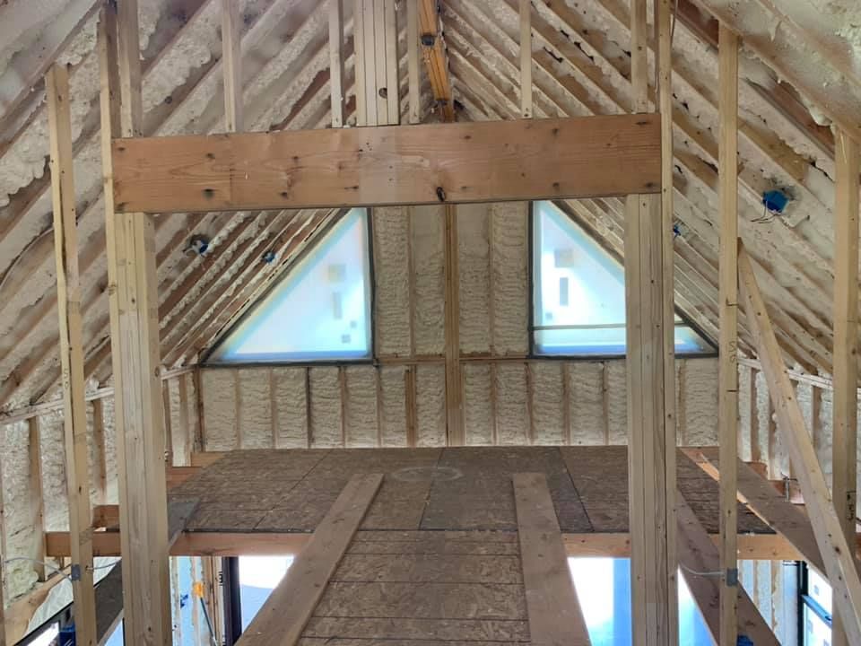 Interior of a framed attic with exposed wooden trusses and small triangular windows at the far end