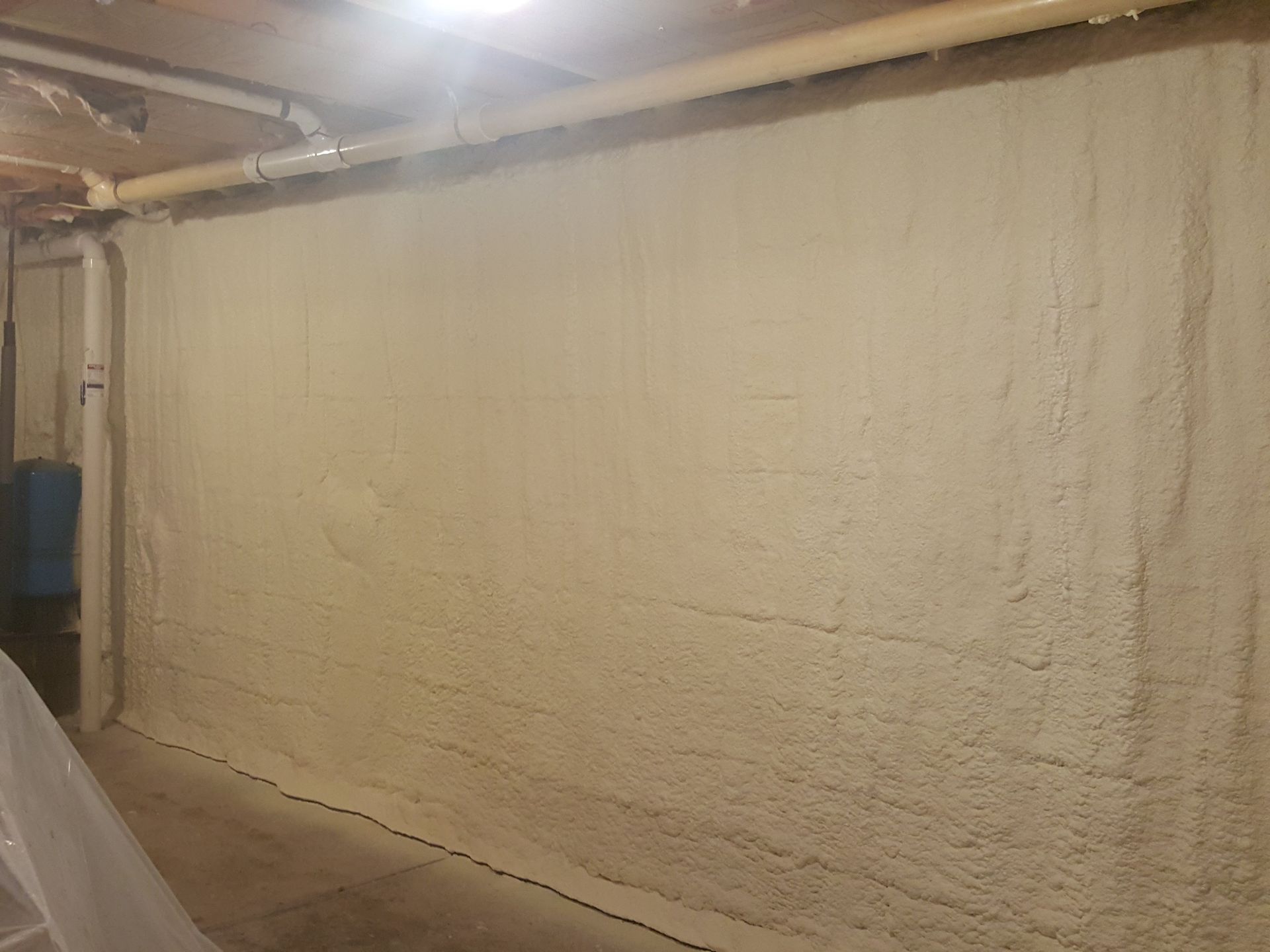 Dim basement corridor with a rough beige wall, exposed pipes, and a concrete floor under bright lights