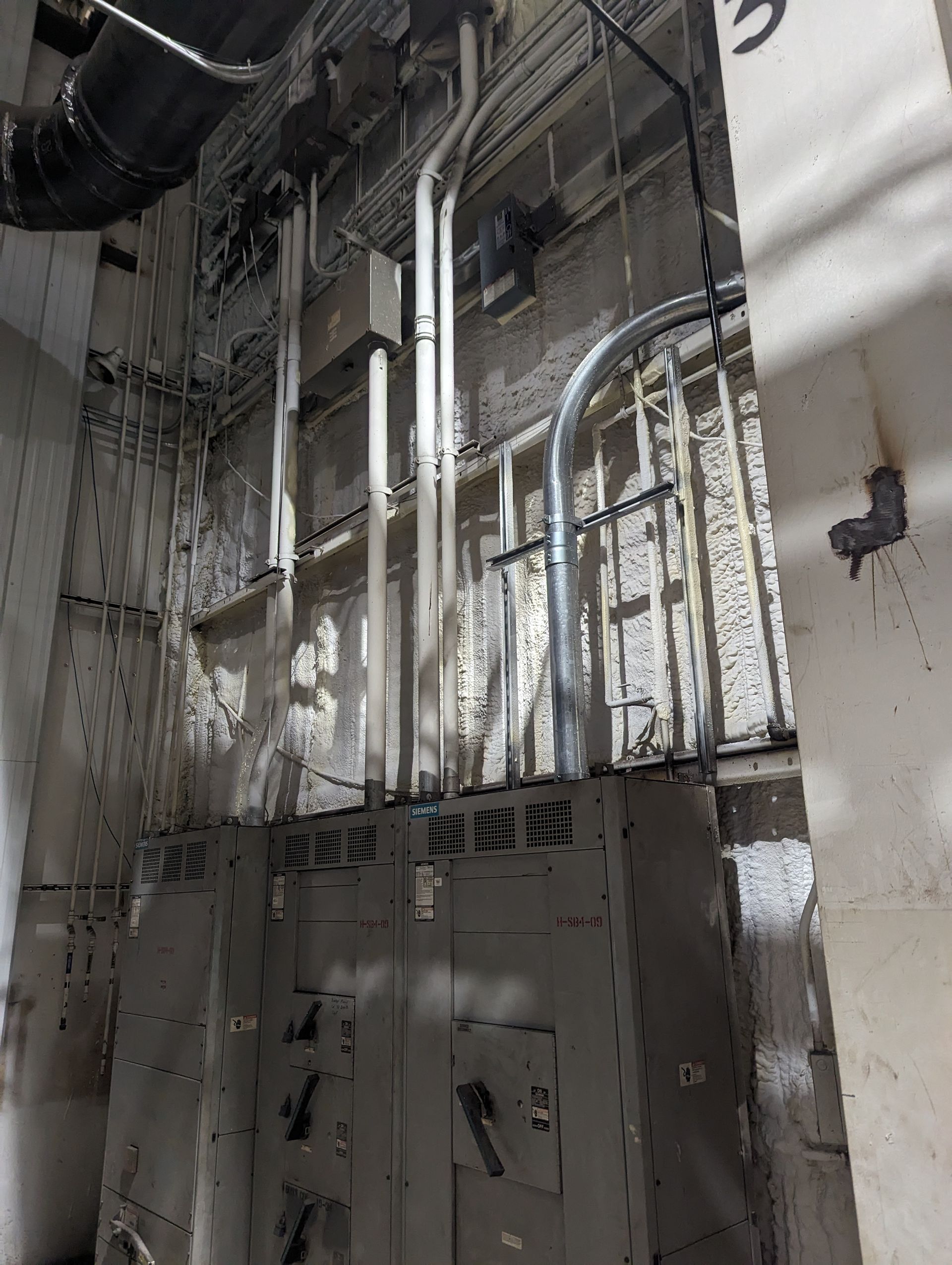 Industrial interior with exposed pipes, ducts, and wiring on unfinished concrete walls.