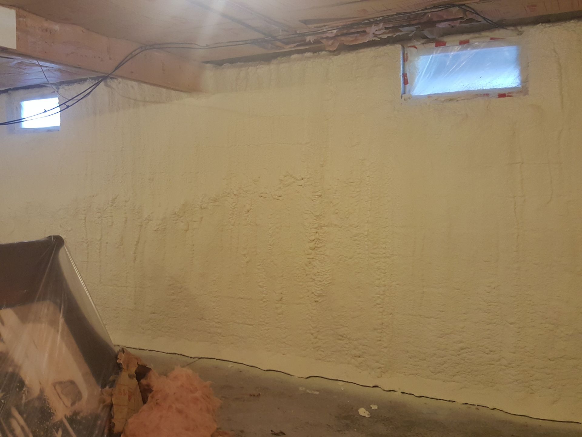 Bare basement wall with two small windows, exposed ceiling beams, and a leaning board in the corner
