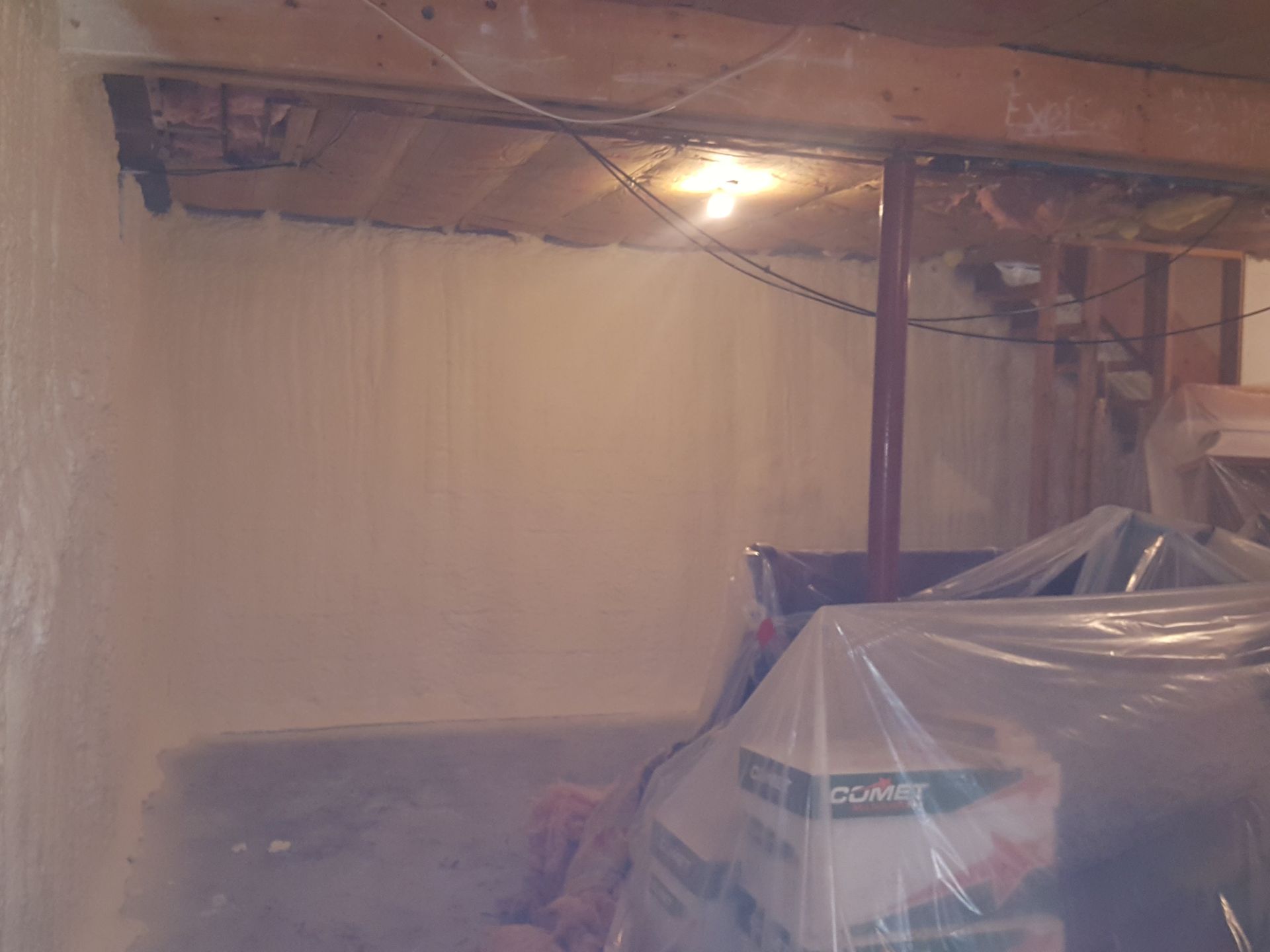 Dim basement with exposed beams, a lightbulb, and a covered furnace wrapped in plastic sheeting.