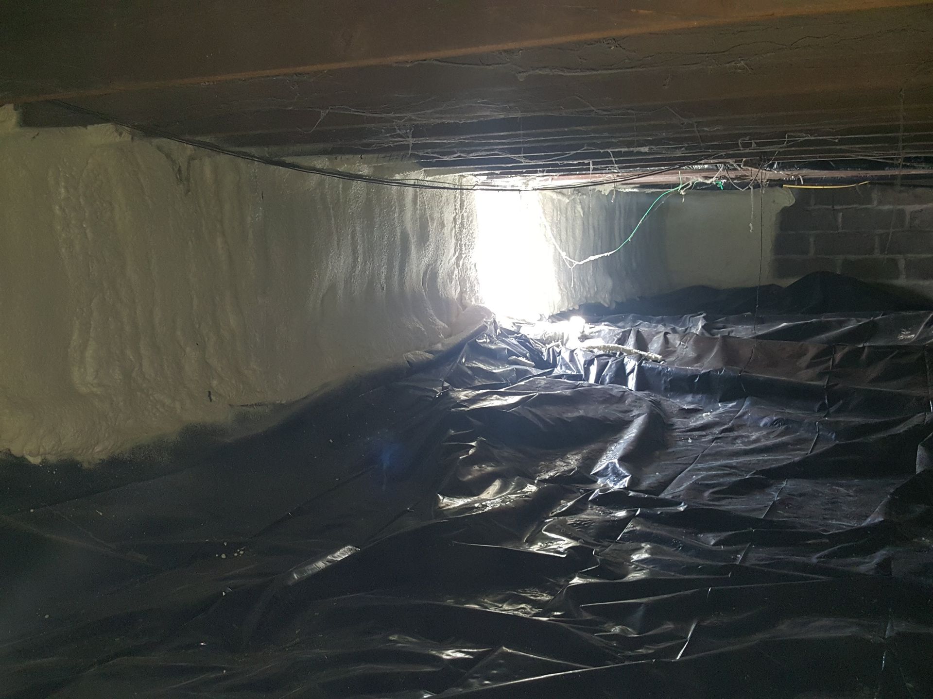 Dark crawlspace lined with black plastic sheeting, lit by daylight from a small opening at one end.