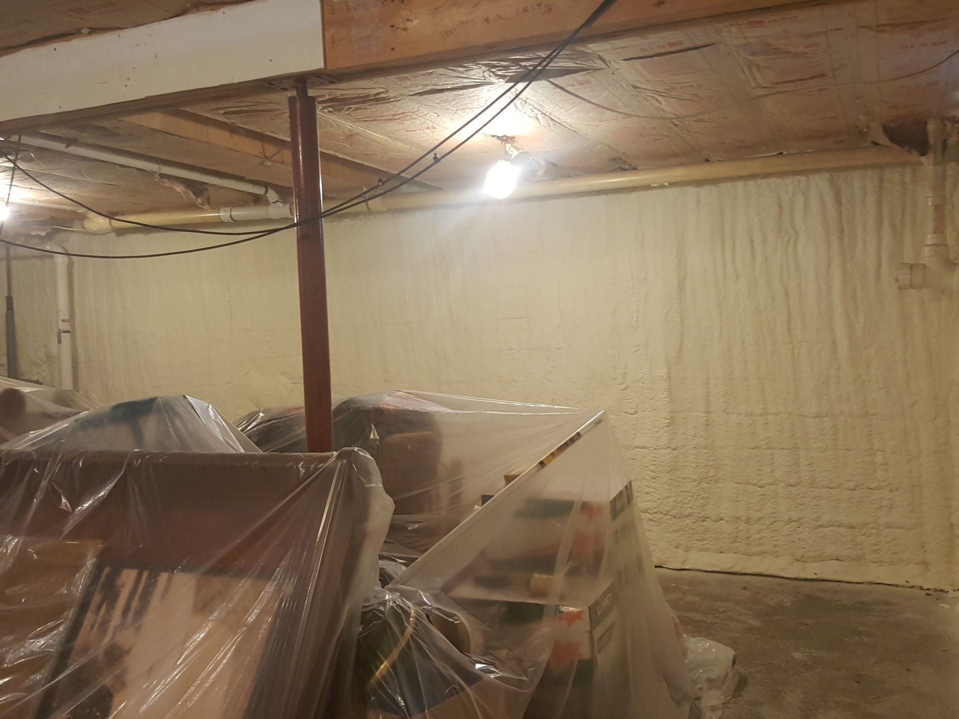 Basement under renovation with plastic-covered furniture, exposed beams, and a bare concrete floor.