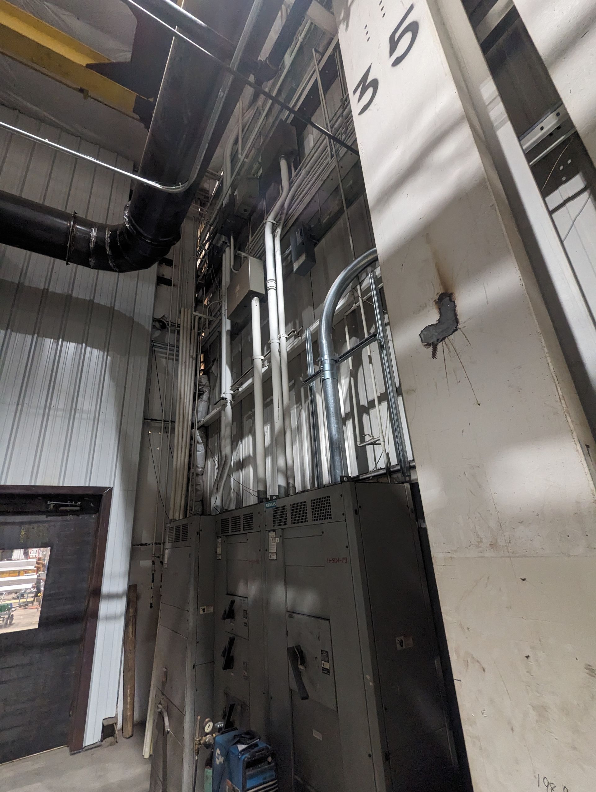 Industrial hallway with exposed pipes, metal panels, and a closed door near the ceiling.