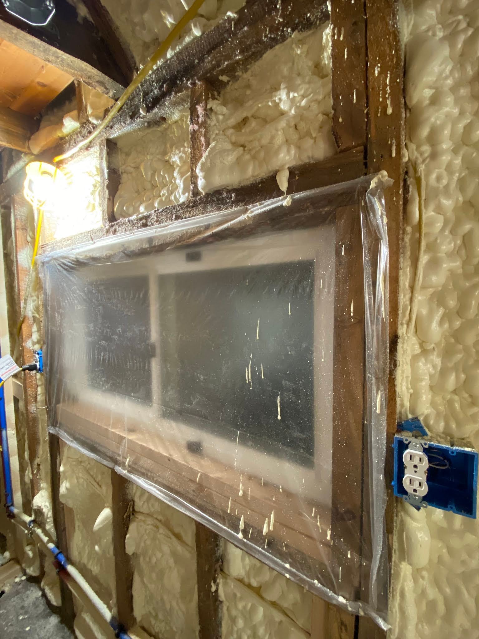 Framed basement window wrapped in plastic, surrounded by exposed insulation and wiring.