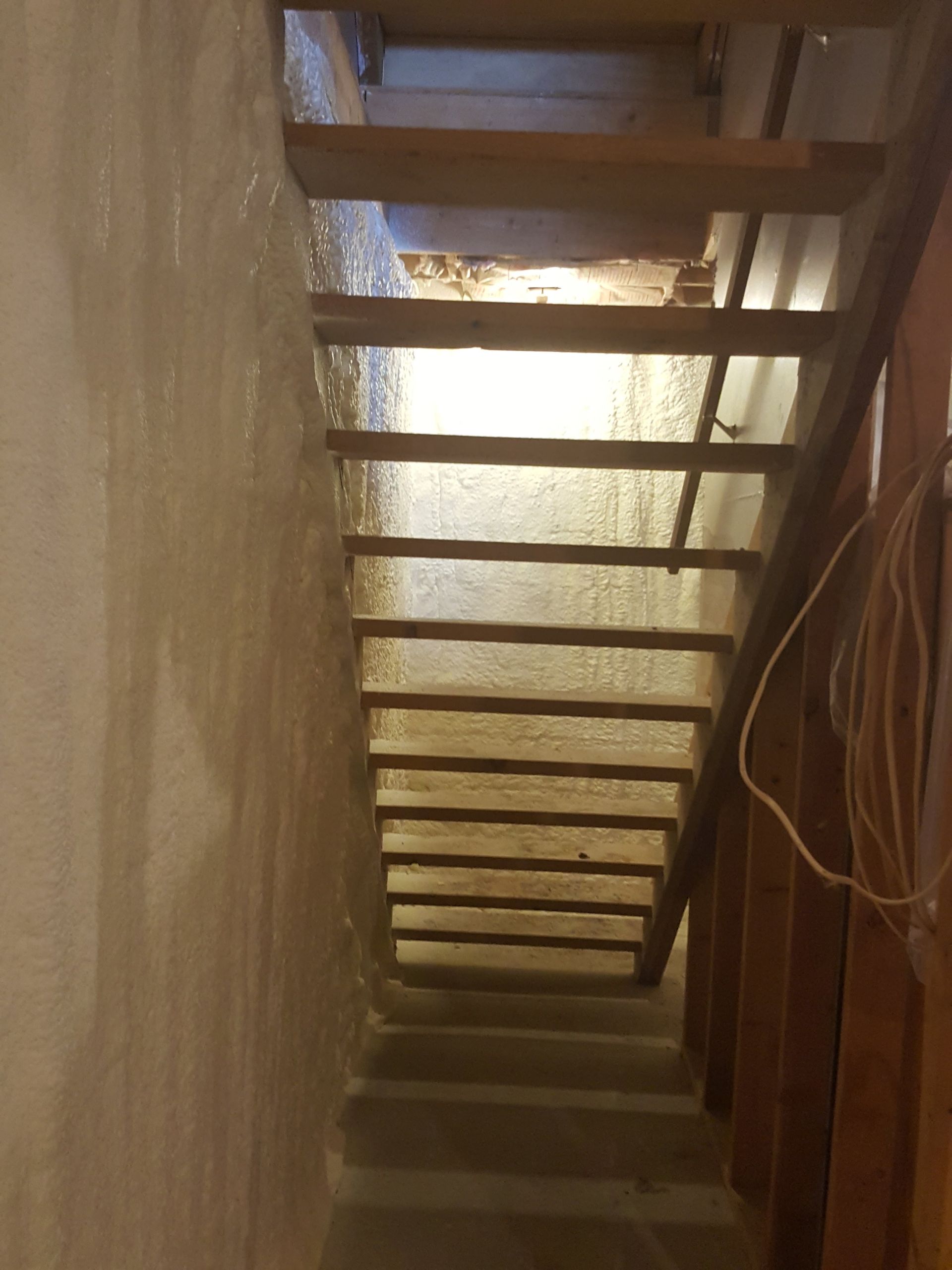 Narrow stairwell viewed upward, with wooden steps and beams framing a dim passage to a bright opening above
