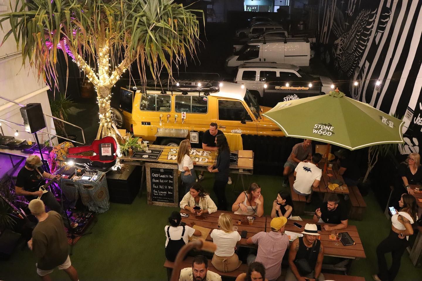Outdoor bar scene with people, picnic tables, food, and music stage. Lit by string lights and a yellow vehicle — Village Bicycle Noosa in Noosa Heads, QLD